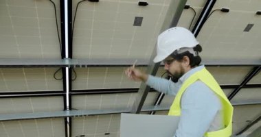 Arka taraftaki güneş panellerini inceleyen adam mühendis ve not defterindeki veri bilgilerini not et. Açık hava güneş enerjisi istasyonunda çalışan kasklı ve üniformalı bir erkek elektrikçi..