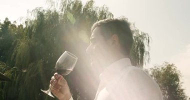 Beyaz gömlekli bir şarap garsonu elinde kırmızı şarapla bardağı tutuyor, aroma kokluyor ve güneşli bir günde açık havada içki tadıyor. Şarap konusunda uzman bir erkek.