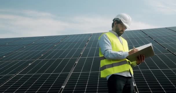 Man Werknemer Uniform Beschermende Helm Staat Fotovoltaïsche Elektriciteitscentrale Onderzoekt Zonnepanelen — Stockvideo