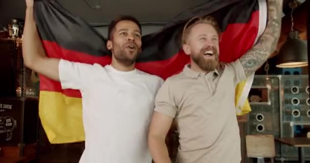 幸せな男性サッカーファンは パブで試合を見ながらドイツの旗を手で振り ジャンプして喜んでいます サッカーのゲームの若い喜びの男性の友人はお気に入りのスポーツチームをサポートします — ストック動画