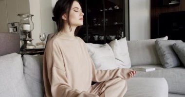 Lotus 'ta oturan kadın gözleri kapalı bir şekilde poz veriyor ve evdeki kanepede meditasyon yapıyor. Kadın oturma odasında farkındalık egzersizi yapıyor. Stres atma konsepti. Ruh sağlığı bakımı