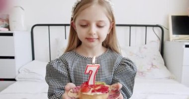 Küçük şirin bir kız doğum gününü kutluyor ve evde pasta mumu üflüyor. Mutlu çocuk yedi yıl kutluyor. Çocukluk konsepti. Çocuk tabağı tatlı tatlı ile süslenmiş yanan mum