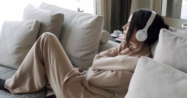 悲しい若い女性は孤独を感じ ワイヤレスヘッドフォンでソファーに横たわり リラクゼーション音楽が自分を抱きしめるのを聞いています ソファーに腰掛けている女性 うつ病コンセプト メランコリームード — ストック動画
