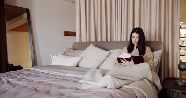 若い焦点を当てた女性はクッションでベッドに横たわり 柔らかい平原に包まれ 休憩中に興味深い本を読みました 寝る前に居心地の良い寝室でリラックスするかなり女性 レジャーコンセプト — ストック動画