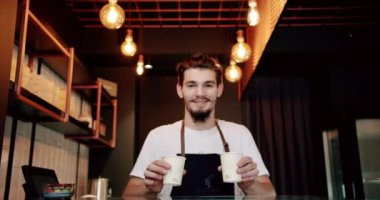 Düşük açılı, beyaz tişörtlü, önlüklü, sakallı bir erkek barista modern kafede kahve servisi yaparken gülümseyip kameraya bakıyor.