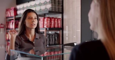 Thoughtful young female customer with long dark hair squinting and smiling while choosing coffee and communicating with crop unrecognizable barista in modern cafe.