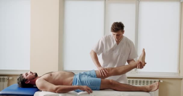 Osteopath在诊所治疗男性患者 骨病门诊康复过程中 男性推拿治疗师伸直无上衣男子腿躺在推拿台上的手持照 — 图库视频影像
