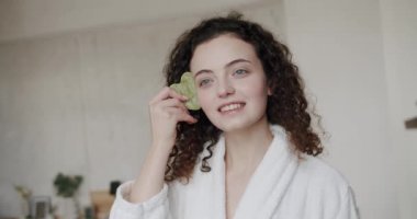 Yeşil bir Gua Sha taş kazıyıcısıyla yüz masajı yapan genç bir kadının portresi. Yüz bakımı ve güzellik ayinleri. Kadın anti-reşit yüz masajı için gua sha yeşim kazıyıcı kullanıyor..