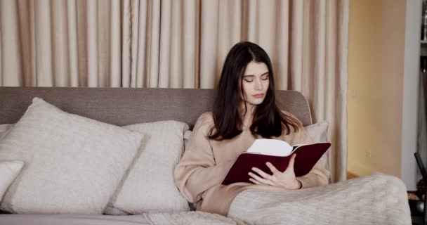 女性はベッドでリラックスし 現代の家の寝室で本を読んでいます 美しい女性は居心地の良いベッドルームで自宅で休んでリラックスしています リラックスして教育の概念のための時間 — ストック動画