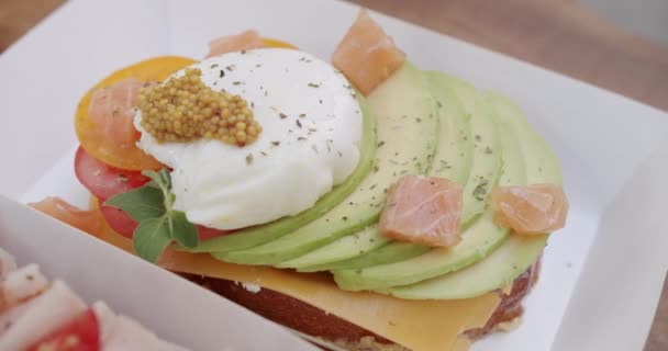 カフェの段ボールパッケージでチーズ アボカドスライス ポーチエッグ サーモン魚の美味しいトーストを閉じます スナックを箱に入れてください 健康的な朝食のコンセプト — ストック動画