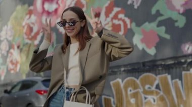 Moda güneş gözlüklü ve kıyafetli güzel kadın sokaklarda barış işareti göstererek ve kamera önünde poz vererek eğleniyor. Pozitif ruh hali olan genç ve şık bir kadın. Blogger yaşam biçimi