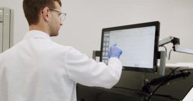 Gözlüklü, üniformalı ve tıbbi eldivenli bir erkek doktor klinikte bilgisayarla çalışıyor. Laboratuvar teknisyeni hastanede profesyonel tıbbi makine kullanarak araştırma yapıyor.