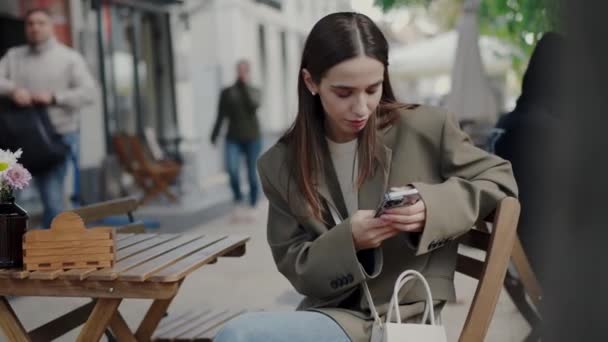 通りのカフェテーブルに座ってスマートフォンでテキストメッセージを送る若いスタイリッシュな女性 屋外の携帯電話でソーシャルメディアを使用してかなり女性チャット コミュニケーションコンセプト — ストック動画