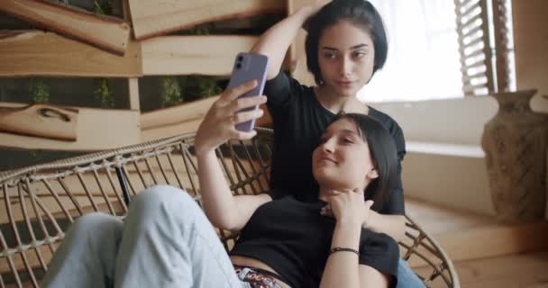 2人の若い女性レズビアンが一緒に時間を過ごし アームチェアでリラックスし スマートフォンでセルフィーを作ります 写真を撮るために携帯電話を使用して幸せな女性カップル — ストック動画