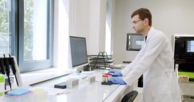 Tıp önlüklü ve gözlüklü bir laboratuvar teknisyeni modern ekipmanlarla hastanede çalışıyor. Erkek doktor kan örneklerini inceleyip klinikte bilgisayar kullanıyor.