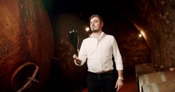 ソムリエは古い地下室でワインを賞賛しています 男性のワインメーカーは ワインセラーで赤ワインを試飲しています ワイン造りのコンセプト 工場でワインボトルを持っている男 多くの木製樽を持つ古いセラー — ストック動画
