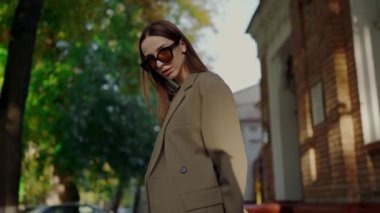 Moda ceketli, güneş gözlüklü, dışarıda poz veren güzel bir kız. Şık giyinmiş ve gözlüklü genç bir kadın şehir sokaklarında yürüyor..