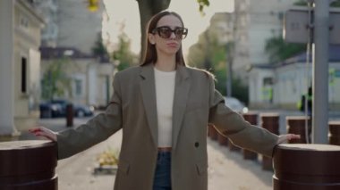 Güneş gözlüklü neşeli bayan şehir caddesinde yürüyor ve yaz günü eğleniyor. Şık giyinmiş, gözlüklü, dans eden ve kamerada poz veren güzel bir kadın.