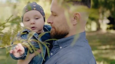 Genç baba küçük oğlunu elinde tutuyor ve yeşil ağaçların yanında parkta yürüyor. Babam tatlı bebekle yaz günü dışarıda vakit geçiriyor. Babalık kavramı