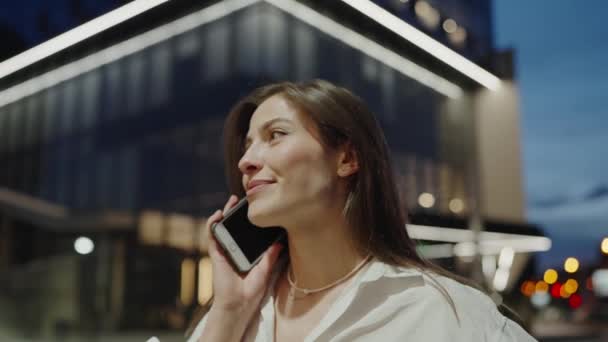 白いシャツの若い女性は 夜に市内のビジネスセンターの近くに立っている間 スマートフォンで話しています かなり女性は仕事の後で屋外の携帯電話で会話を持っています — ストック動画