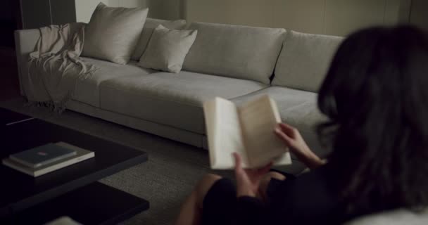 女性は インテリアデザインの設定でスタジオソファーに快適に座って 彼女の太もとに残っている本を読んでいます — ストック動画