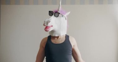 Tek boynuzlu at maskeli ve güneş gözlüklü komik bir adam ağırlıklarla evde beyaz arka planda antrenman yapıyor. Esprili esnemeler. Komik ev egzersizi.