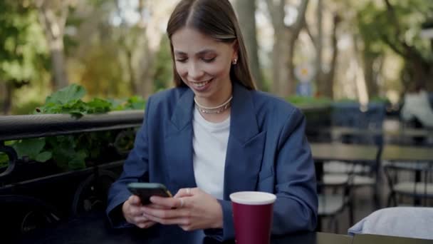 时尚漂亮的女人坐在外面的咖啡桌旁 用智能手机在网上聊天 在餐馆喝咖啡休息时 迷人的女性在手机上滚动社交媒体 微笑着 — 图库视频影像