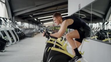 Adam spor salonunda egzersiz bisikletine biniyor, şort giyiyor ve bacaklarıyla otomotiv tekerleği üzerinde çalışıyor, asfalt üzerinde bir ata biniyormuş gibi yapıyor.