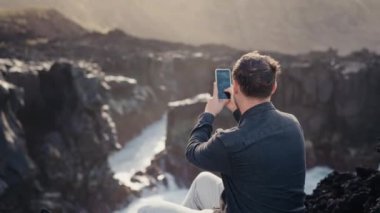 Bir adamın arka görüntüsü kayalık sahil kıyısında akıllı telefon kullanarak su sıçratan denizin videosunu çeker. Cep telefonlu bir erkek okyanus kıyısında güzel manzaralı bir uçurumda oturuyor.