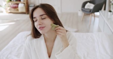 Yeşil bir Gua Sha taş kazıyıcısıyla yüz masajı yapan genç bir kadının portresi. Cilt kozmetolojisi kavramı. Doğal güzellik estetiği. Evde güzellik tedavisi..
