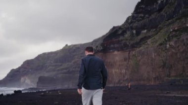 Genç adam deniz kıyısında yürüyor ve dağlara bakıyor güzel doğanın tadını çıkarıyor. Rüzgârlı bir havada okyanus kıyısındaki bir erkeğin arka görüntüsü. Seyahat ve macera konsepti