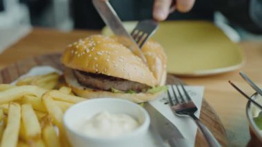 Kafe masasında otururken çatal ve bıçak kullanarak lezzetli hamburgerleri etle kesen erkek ellerine yakın çekim. Lezzetli hamburger ve soslu patates kızartması yiyen aç bir adam. 