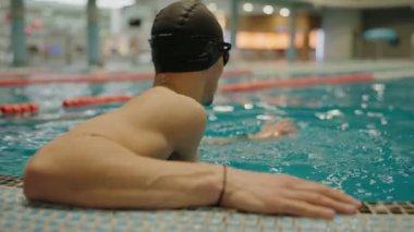 Yüzme şapkalı ve gözlüklü bir adam yüzme havuzu eğitimi arasında mola verir ve dinlenir. Profesyonel sporcu yüzme havuzunun suyunda egzersiz yapıyor.