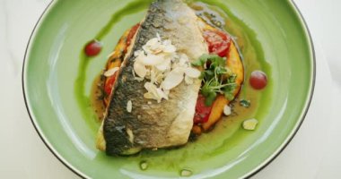 Patates püresi ve sebzeli güzel dekore edilmiş beyaz balık parçası yeşil tabakta ve beyaz arka planda dönüyor. Yavaş çekim deniz levreği, bbq sebzeleriyle servis edilir. Yakın çekim. Üst Görünüm.