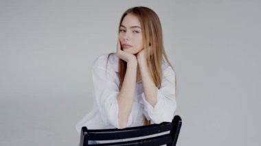 Katmanlı saçlı, beyaz gömlekli bir kadın siyah bir sandalyede oturuyor, kolu koltuklarda dinleniyor ve elleri kalçasında dinleniyor.