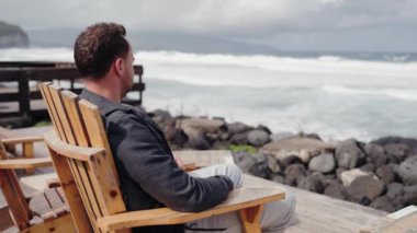 Terasta tahta koltukta oturan ve okyanusu izleyen bir adam. Erkek doğada dinleniyor ve deniz manzarasının tadını çıkarıyor. Tatil kavramı