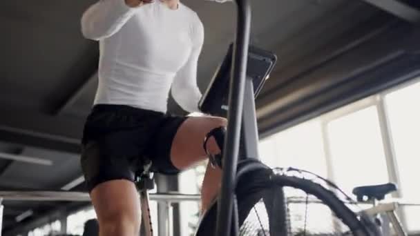 男性はジムでエクササイズマシンをサイクリングし ショートパンツを着用しながら太もももももひもを働いています 自転車フレームに取り付けられた車輪に取り付けられた自転車タイヤにペダルをかけています — ストック動画