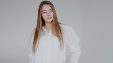 Uzun saçlı, beyaz gömlekli, kendine güvenen bir manken beyaz arka plan önünde poz veriyor, elleri ceplerinde ve saçlarını tutarak güçlü bir göz teması kuruyor.