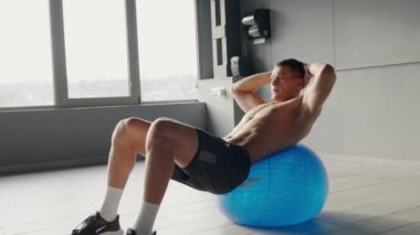 Bir adam spor salonundaki mavi bir egzersiz topunun üzerinde rahatça uzanır, dizini ve bacağını dinlendirirken,