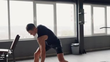 Bir adam spor salonunda halter kaldırıyor, dirseği bükülürken göğsünü, kalçasını ve dizini esnetiyor. Binanın ahşap döşemesi insan bacağı için boş bir aktivite sağlıyor.