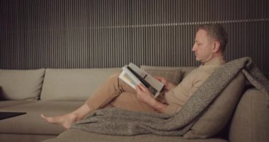 Hafta sonları evde dinlenirken rahat koltukta oturup dergi okuyan odaklanmış bir adam. Erkek, evde, kanepede dinleniyor.