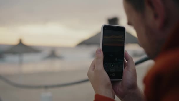 スマートフォンを持っている男と電話カメラを使用して海岸の撮影ビデオに選択的な焦点 男性の散歩や休暇中に携帯電話で海の景色で美しい自然の写真を作る — ストック動画