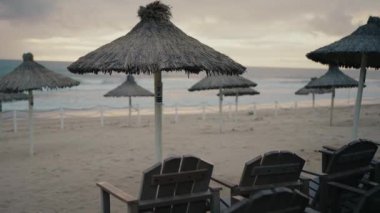 Tahta rahat sandalyeler ve güneş batarken okyanus kumlu sahilinde saman plaj şemsiyeleri. Deniz kıyısında boş şezlonglar ve güneş şemsiyeleri var. Tatil kavramı