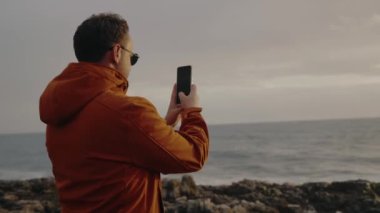 Arkaya bakan adam akıllı telefonu tut ve güzel deniz manzarasının fotoğrafını çek. Okyanus kıyısında yürüyen ve cep telefonu kamerasıyla doğanın videosunu çeken genç bir erkek.
