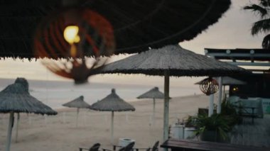 Açık hava kafesi, dekoratif lambalı saman şemsiyeler, tahta sandalyeler ve yazın okyanusun yanındaki kumlu sahilde masa. Sabahın erken saatlerinde güneşli güneş şemsiyeleri ve şezlonglarıyla bomboş deniz kıyıları