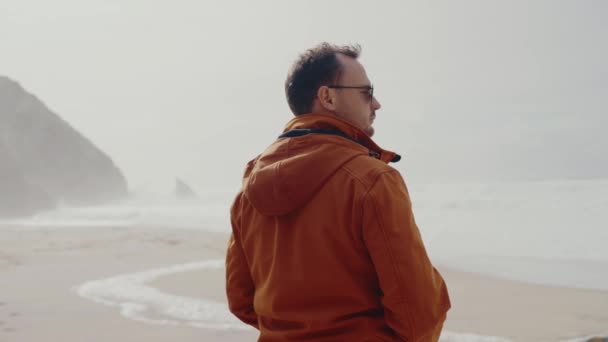 海岸に立っているサングラスの男性と 朝の波の水を見ている男性の背中を見てください 男性は屋外で海岸を歩き 休暇や週末に孤独を楽しんでいる — ストック動画