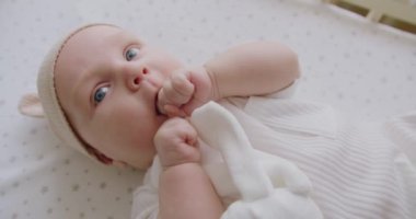 Tombul yanakları ve büyük mavi gözleri olan sevimli yeni doğmuş çocuk vücudunu hareket ettiriyor ve kameraya bakıyor. Güzel, yeni doğmuş bebek yatak çarşaflarında yatıyor, ayak parmaklarına dokunuyor gülüyor kameraya bakıyor..
