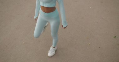 Mavi spor giysili, spor giyinmiş, bacaklarını açık havada açan genç bir kadın. İnce vücutlu, aktif giyimli ve spor ayakkabılı bir kız antrenman için ısınıyor. Moda spor kıyafeti.