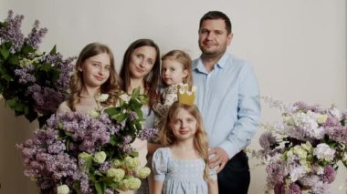 Stüdyoda çiçekli beyaz bir arka planda fotoğraf için duran aile portresi. Ağır çekimde mutlu bir ailenin portresi. Çocuklarla birlikte mutlu genç bir aile..