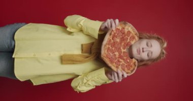 Sarı tişörtlü, biberli pizza yiyen, kırmızı izole edilmiş arka planda eğlenen ve gülümseyen güzel aç kızın dikey videosu. Lezzetli pizza yiyen kadın pepperoni büyük dilim.